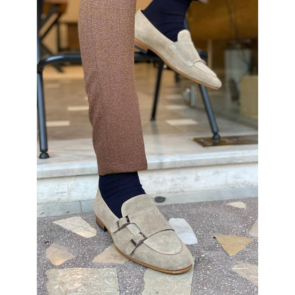 Close-up of a man wearing beige suede double monk strap loafers with navy socks and brown textured trousers, standing near a marble step.