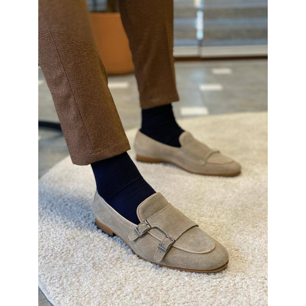 Close-up of a man wearing beige suede double monk strap loafers with navy socks and brown textured trousers, standing near a marble step.
